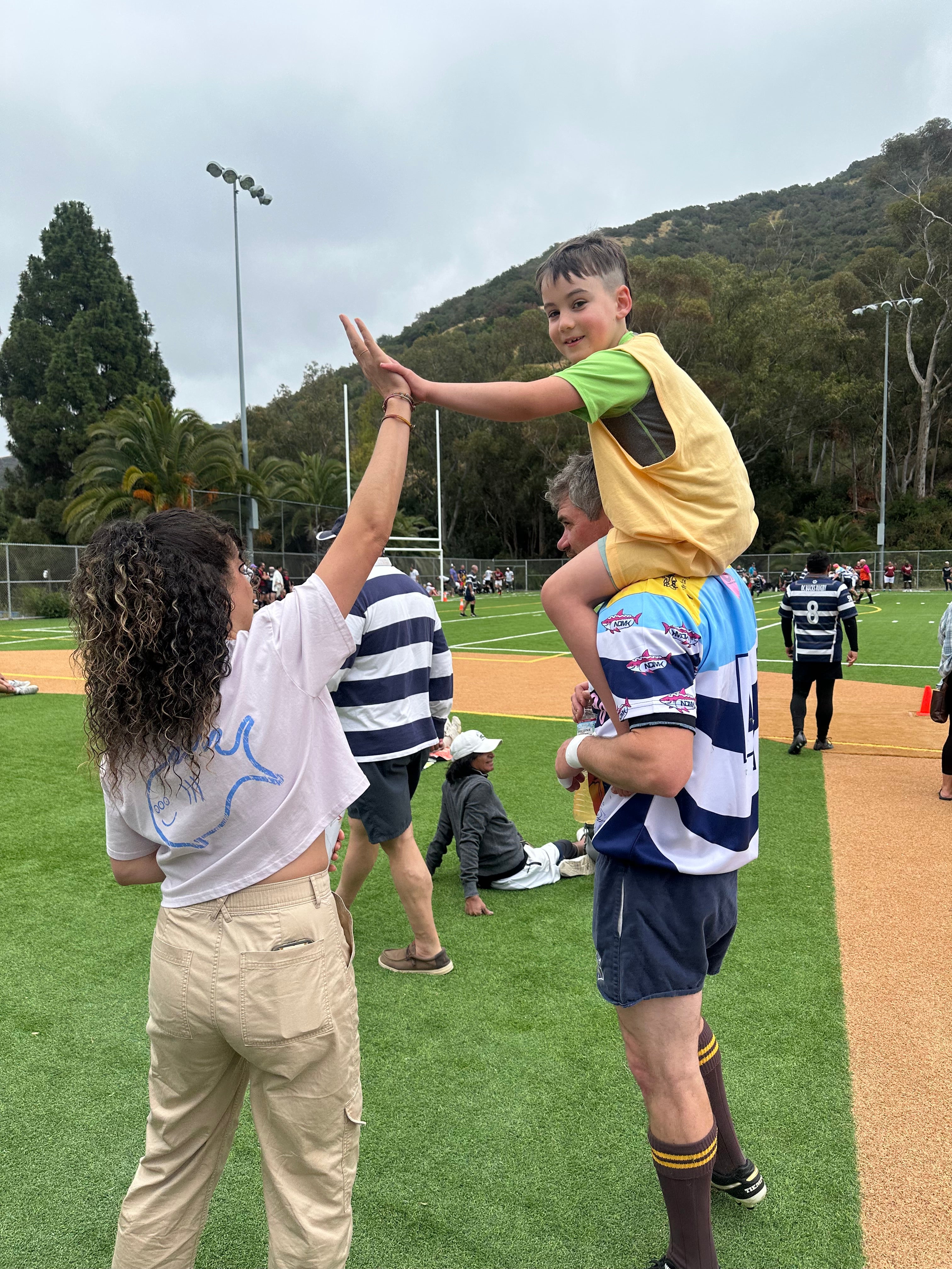 A man wearing a blue and white striped rugby jersey carries a young boy in a yellow bib while high-fiving a woman on a sports field.