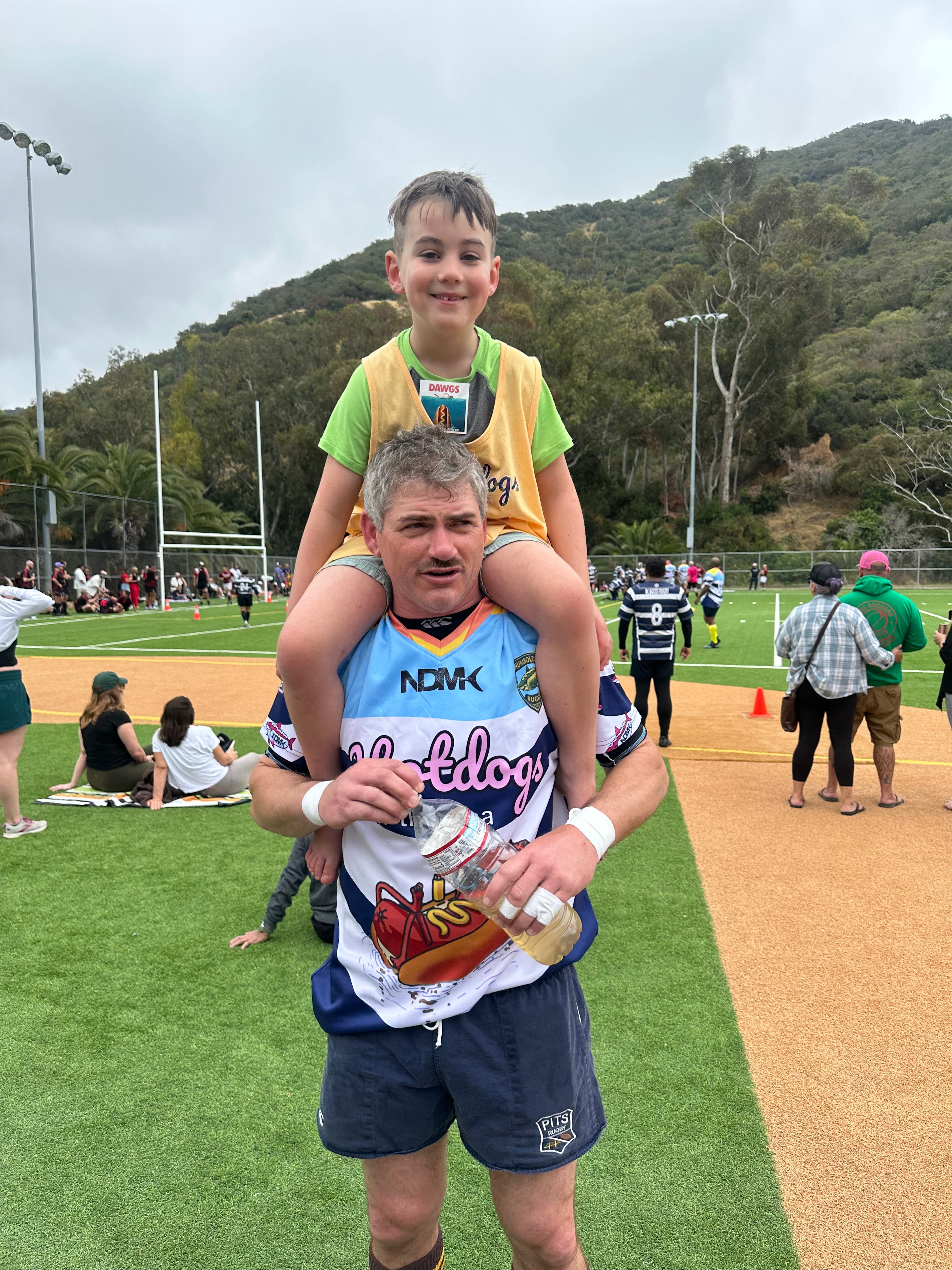 A man on a sports field wearing a blue and white Bulldogs jersey with 'NSW' and 'Bulldogs' visible, carrying a young boy on his shoulders. The boy is wearing a yellow athletic bib.