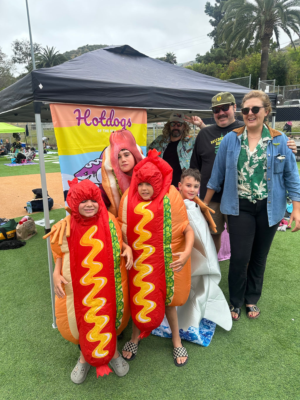 A group of five people posing outdoors in front of a black canopy tent on artificial turf. Two children are wearing oversized hot dog costumes, while the adults wear casual outfits.